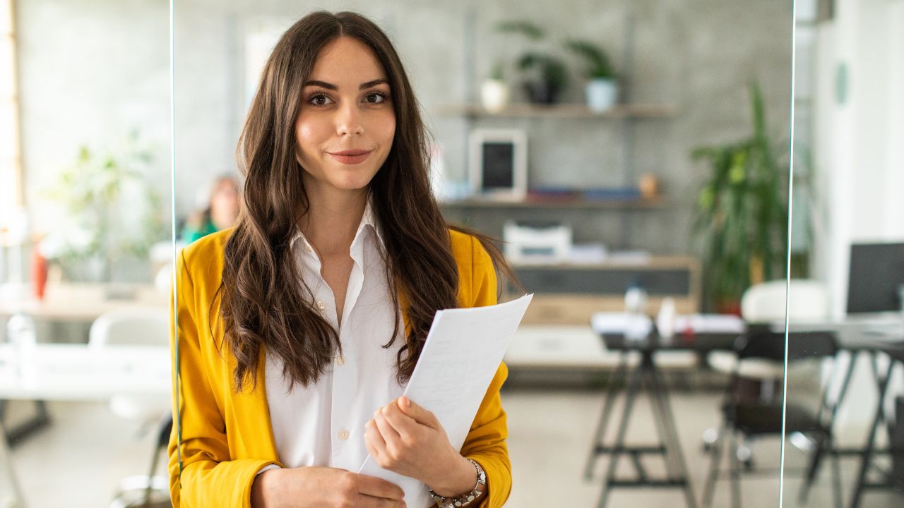 A business woman smiling at the camera while standing in an office, holding a folder