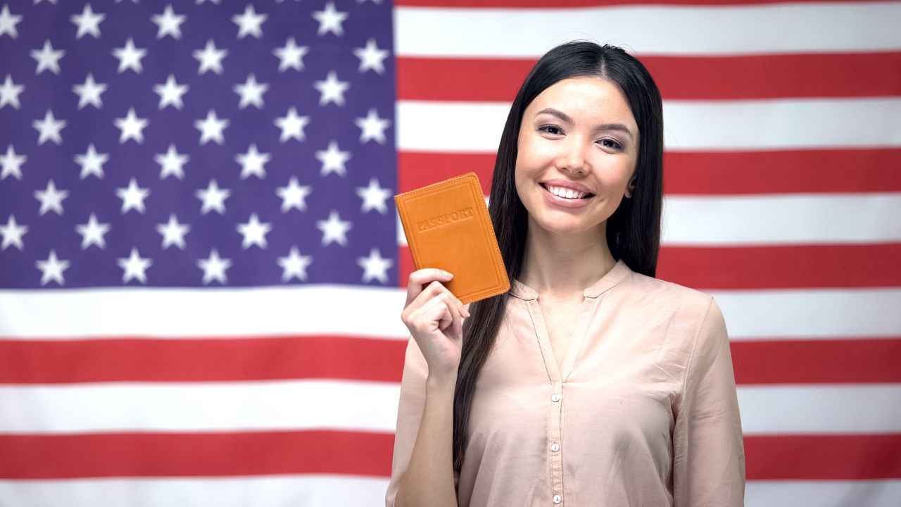 A woman smiling in front of a US flag while holding a passport