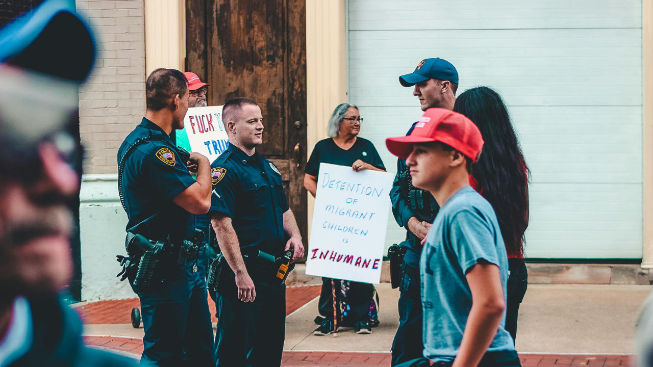 Protesters and police officers at a rally with signs about immigrant detention and deportation.