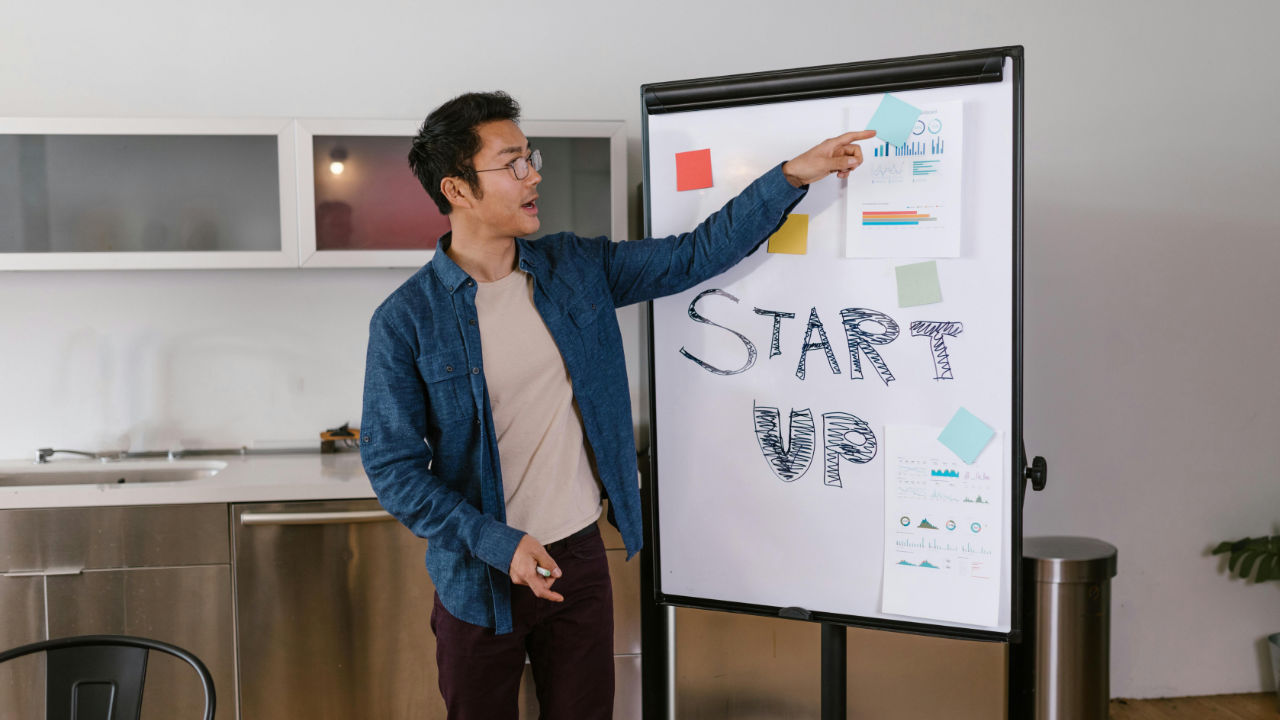 A man in a blue shirt and glasses points to charts on a whiteboard that has the words "START UP" written on it.