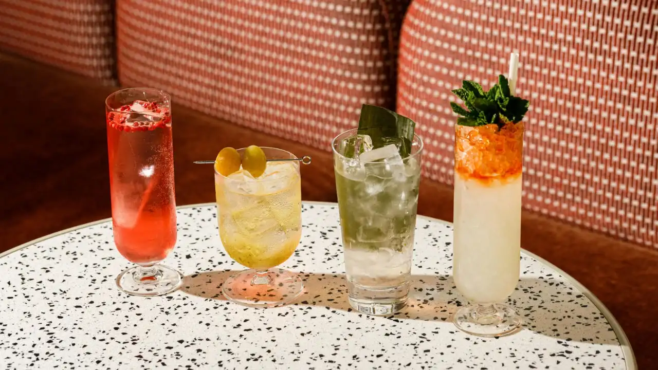 Four different craft cocktails in various glasses sit on a white speckled table in front of a red patterned booth.