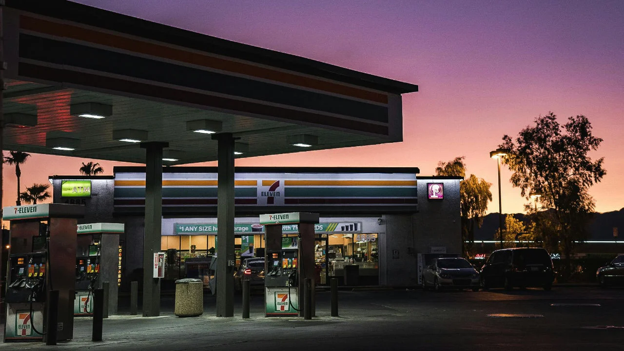 An illuminated gas station and 7-Eleven convenience store against a purple and orange sunset sky.