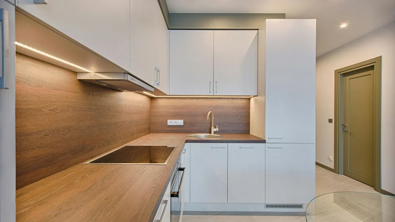 A modern kitchen with clean lines, featuring white cabinets, wood-grain countertops and backsplash, and under-cabinet lighting.