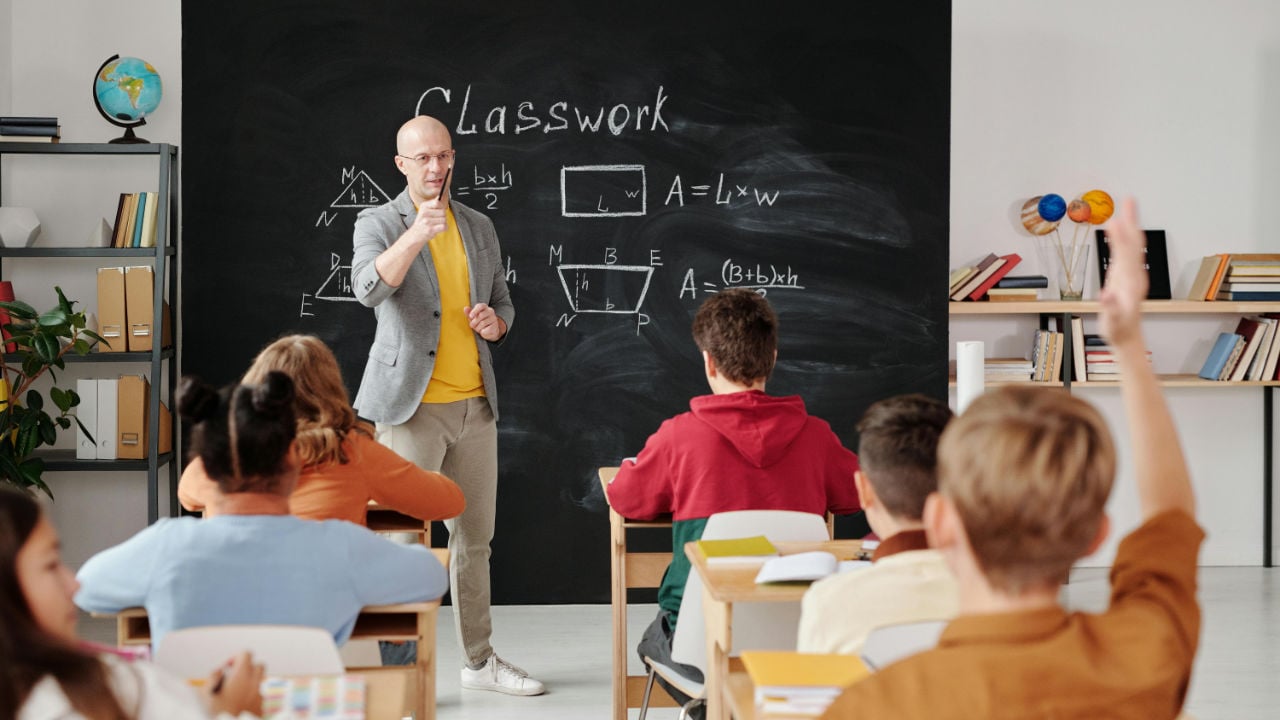 A male teacher in a grey blazer points to students in a classroom, with a chalkboard covered in math formulas behind him.