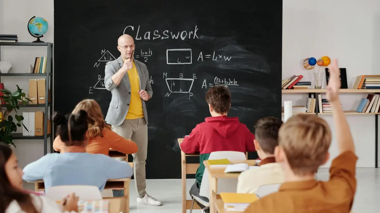 A male teacher in a grey blazer points to students in a classroom, with a chalkboard covered in math formulas behind him.