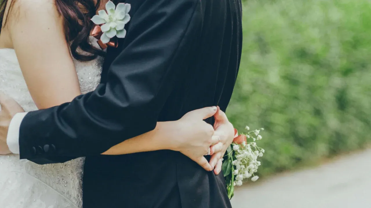 A close-up of a bride in a white lace dress and a groom in a black suit hugging each other. The bride is holding a small bouquet.
