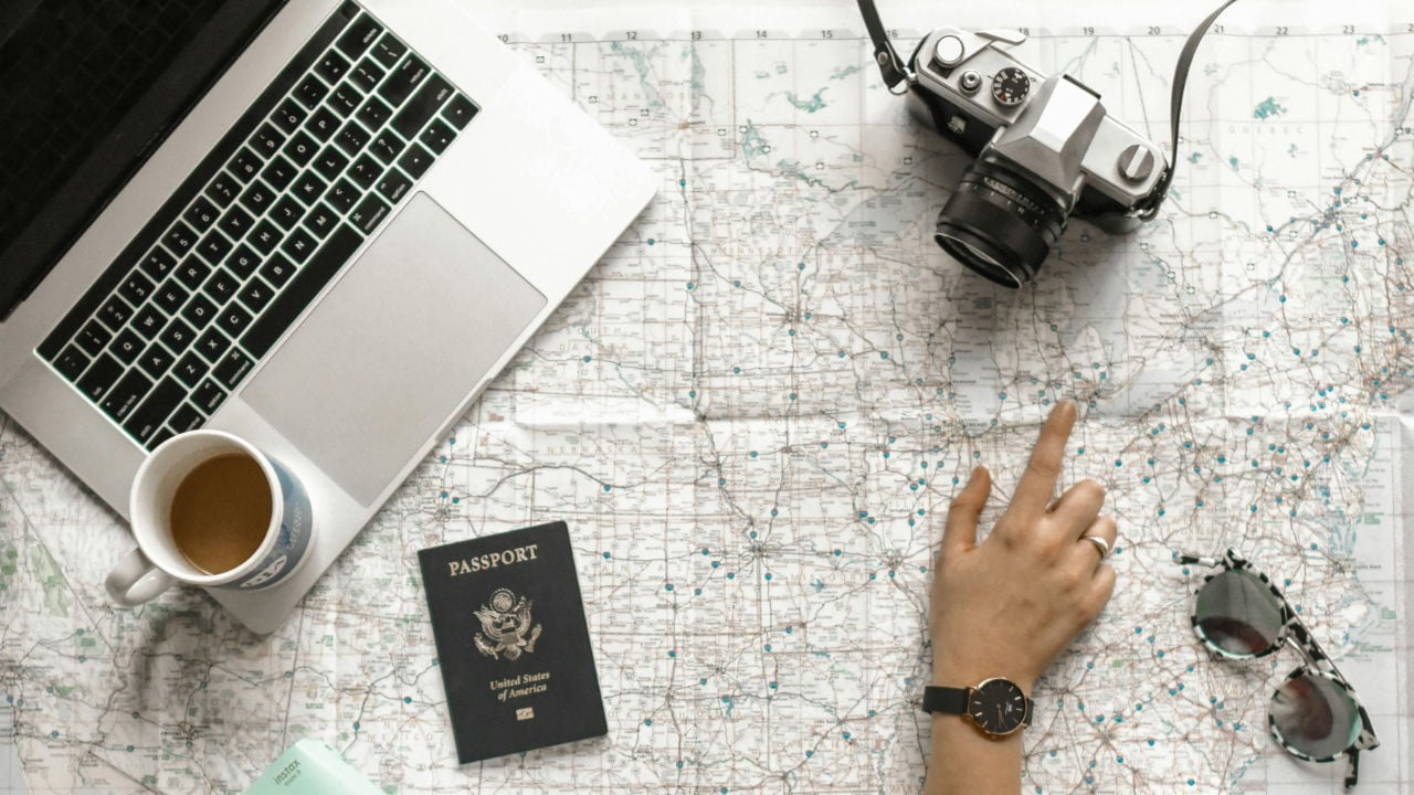 An overhead view of a person's hand pointing at a large map, surrounded by a laptop, a U.S. passport, a camera, and a cup of coffee.