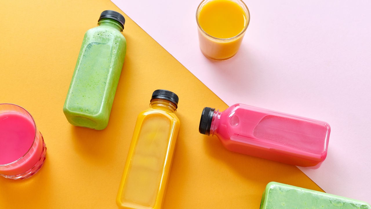 An arrangement of different colored juices in clear bottles and glasses on a bright orange and pink background.