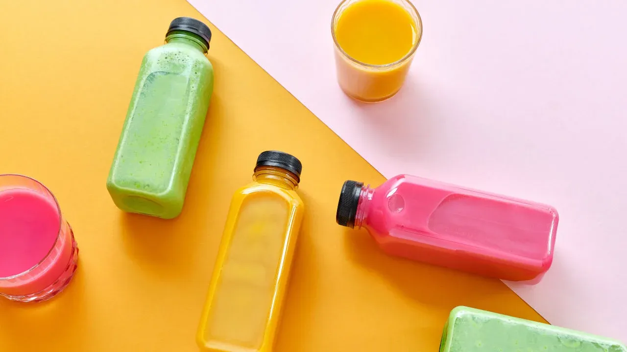 An arrangement of different colored juices in clear bottles and glasses on a bright orange and pink background.