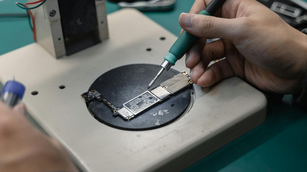 A close-up of a person's hands using a small screwdriver to work on a microchip under a magnifying lamp.