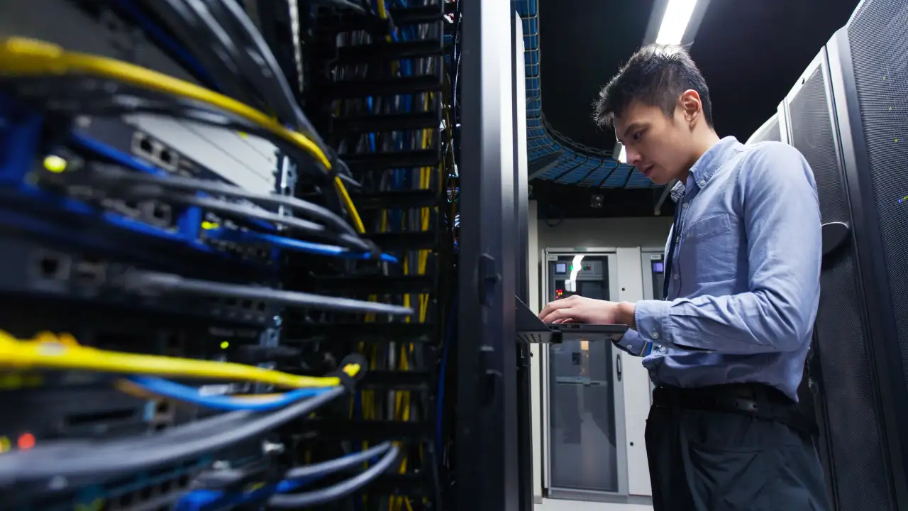 A young professional in a blue collared shirt works on a laptop in a server room, surrounded by complex racks of computer servers and colorful cables.