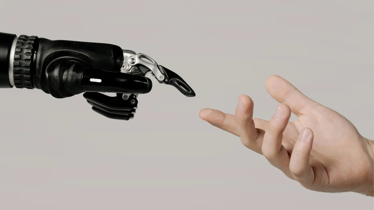 A black and silver robotic hand reaches out to touch the fingers of a human hand against a plain grey background.