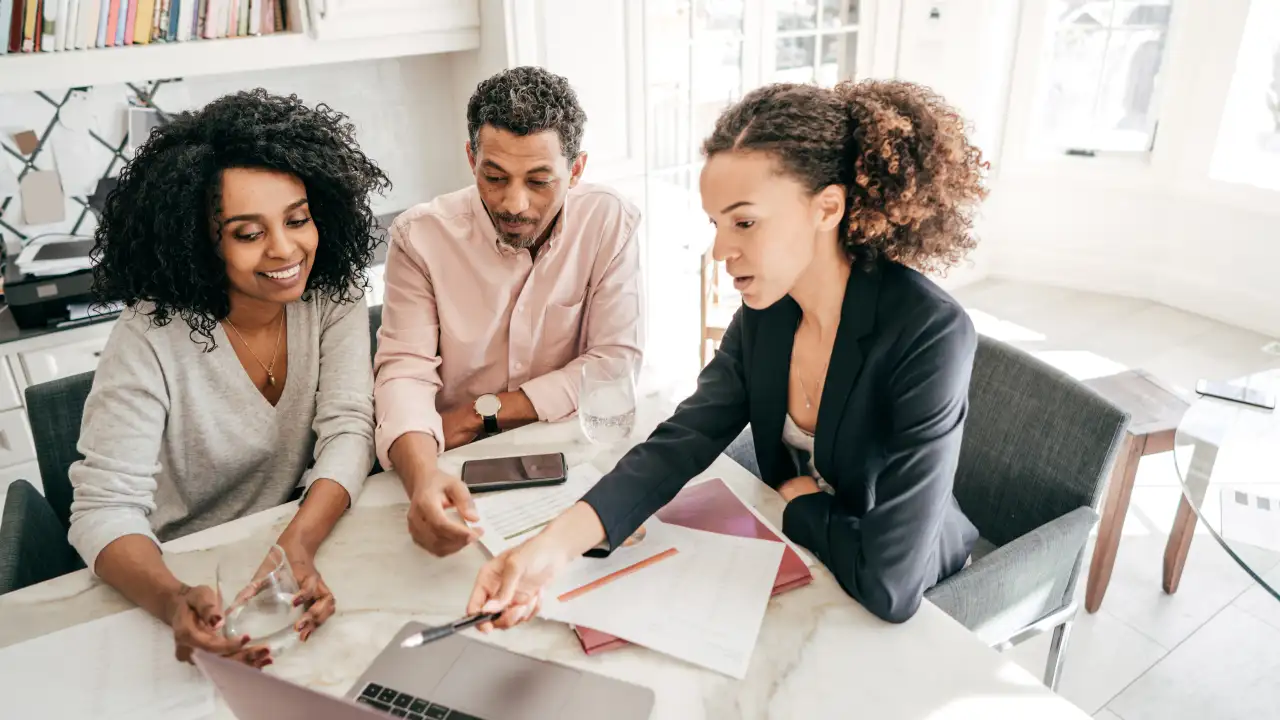 A legal or business professional, a woman in a black jacket, points to a document on a laptop screen while consulting with a male and female couple in a brightly lit kitchen.