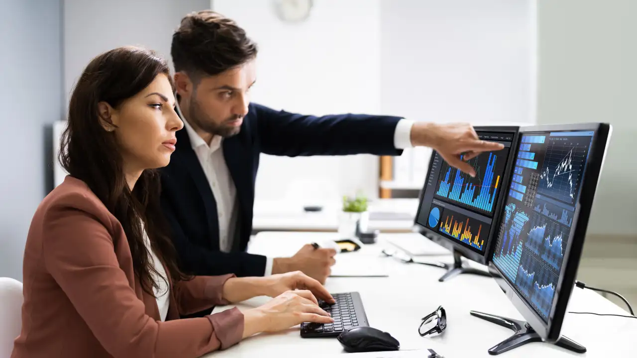 A man and a woman in professional attire looking at charts and graphs on multiple computer monitors in an office setting. The man points to a graph on a screen while the woman types on a keyboard.