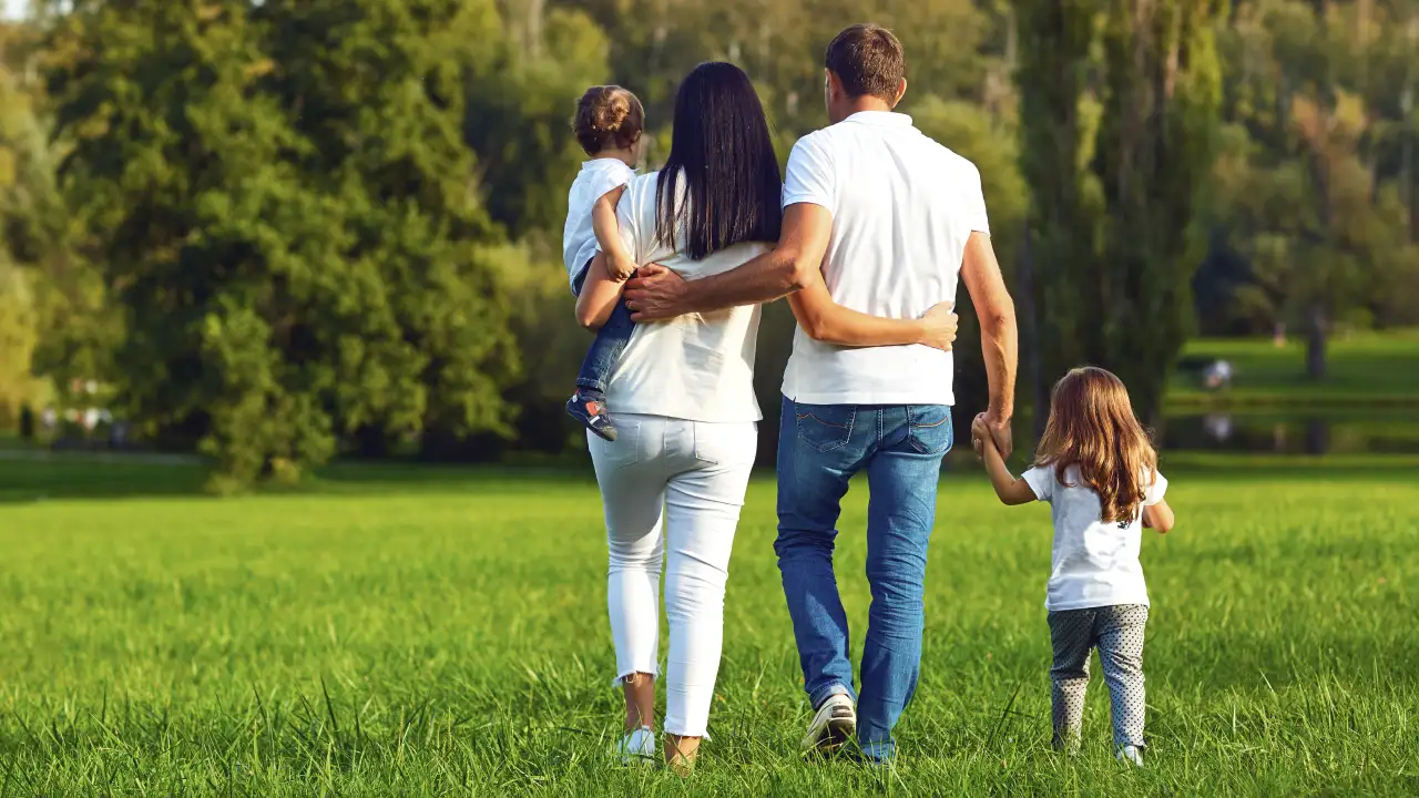 A family of four, including a mother, father, and two young children, walks hand-in-hand through a grassy field, with their backs to the camera, representing a secure future.