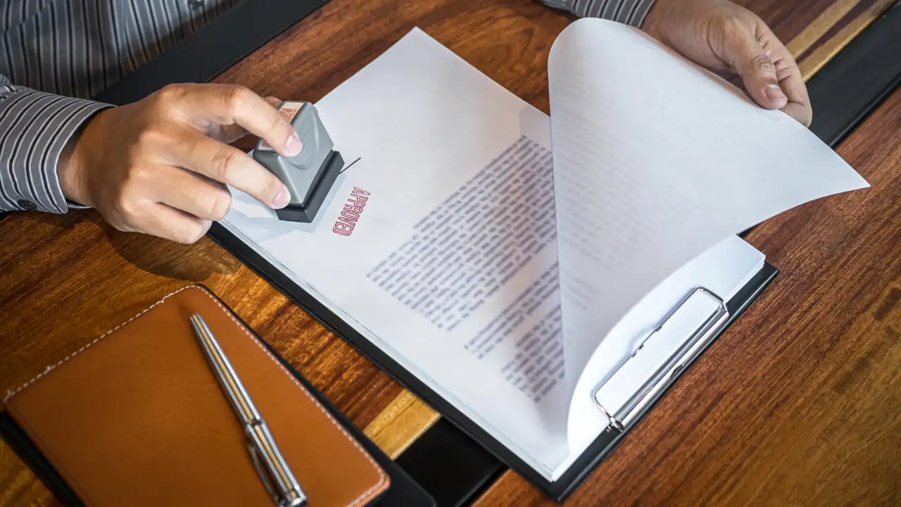 A person's hands stamping a document on a clipboard with a red 'APPROVED' stamp on a wooden desk.