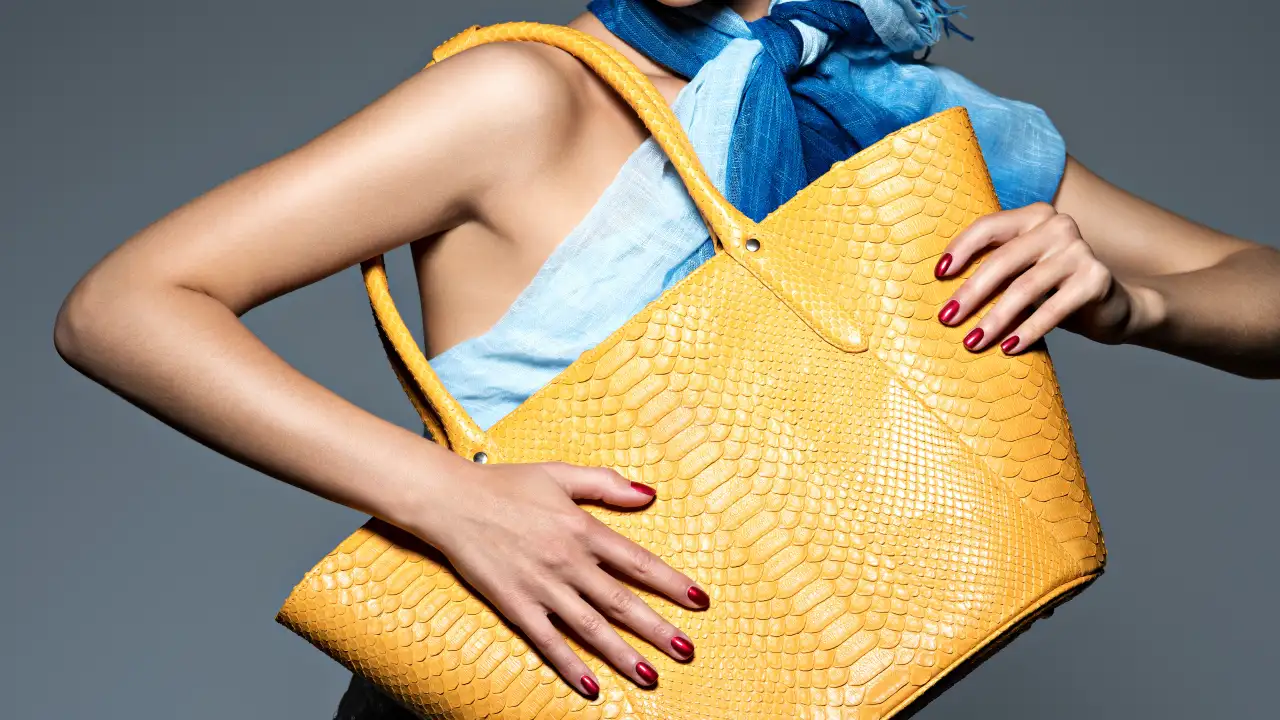 A close-up shot of a woman with red nail polish holding a large, textured yellow luxury handbag over her shoulder, with a blue patterned scarf visible.