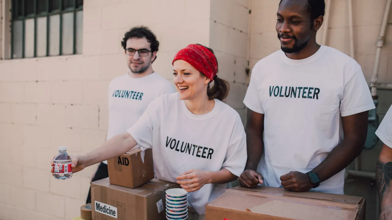 A diverse group of people wearing white "VOLUNTEER" t-shirts smile while working at a table with boxes labeled "AID" and "Medicine," representing a non-profit organization's charitable mission.
