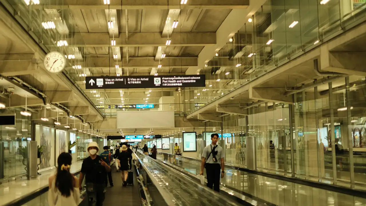 A view inside a modern airport terminal with a moving walkway, glass walls, and overhead signs indicating "Visa on Arrival" and immigration checkpoints.