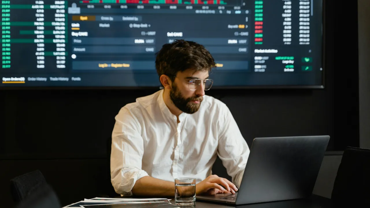 A professional man at a laptop analyzing financial data on a large screen, representing a TN Economist applicant.