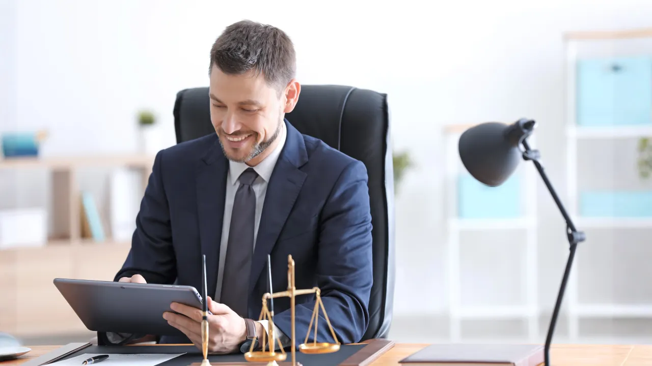 A middle-aged man in a suit and tie, who appears to be an attorney, sits at a desk in a bright office, happily looking at a tablet with a scale of justice figurine nearby.