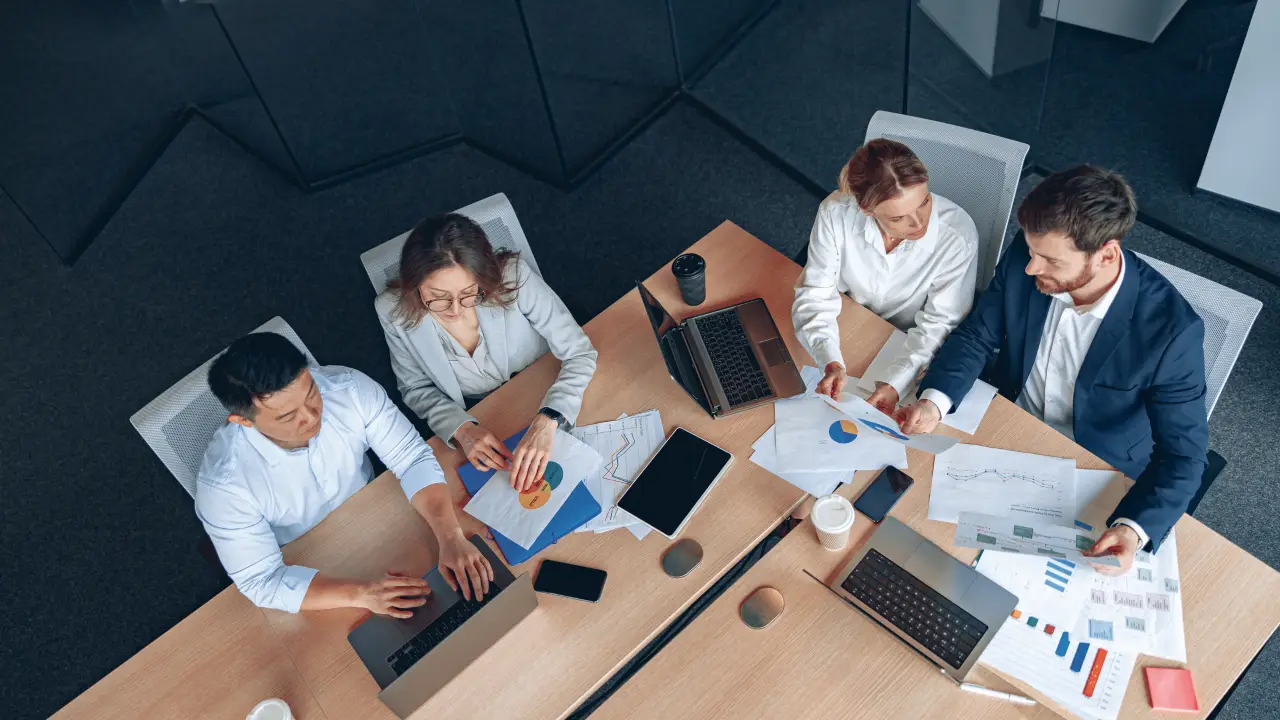An overhead shot of four business professionals sitting at a conference table with laptops, papers, and charts, discussing data and financial graphs.