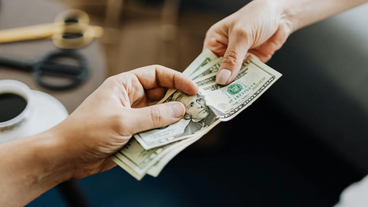 A close-up of two pairs of hands exchanging US dollar bills, with only the hands and money in focus.
