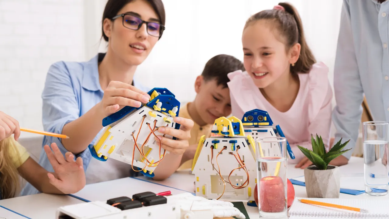 A female educator wearing glasses helps a group of diverse young students assemble small, modular robots in a bright classroom setting, with smiles on the children's faces.