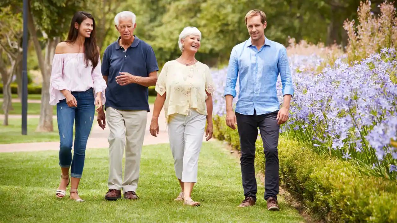 A multi-generational family of four—two older adults and two younger adults—smiling and walking on a grassy path next to a colorful row of purple flowers in a park.