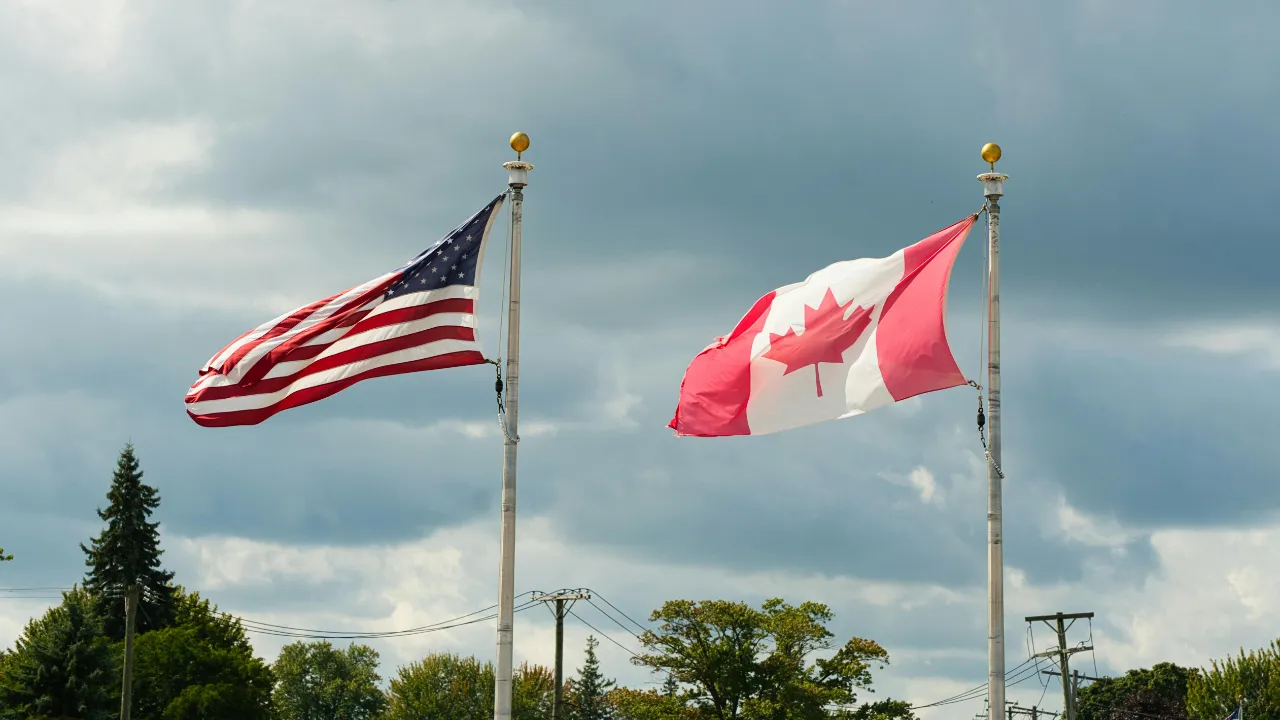 American and Canadian flags flying side by side on flagpoles under a cloudy sky with trees and rooftops in the background.