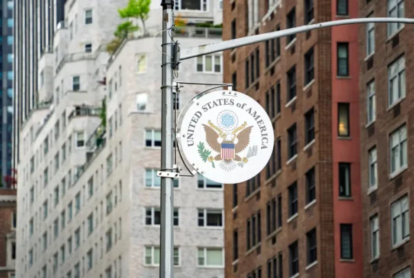 A street sign featuring the emblem of the U.S. government in focus, with blurred city buildings in the background.