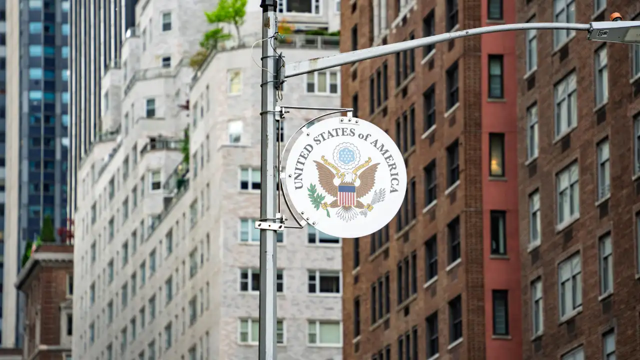 A street sign featuring the emblem of the U.S. government in focus, with blurred city buildings in the background.