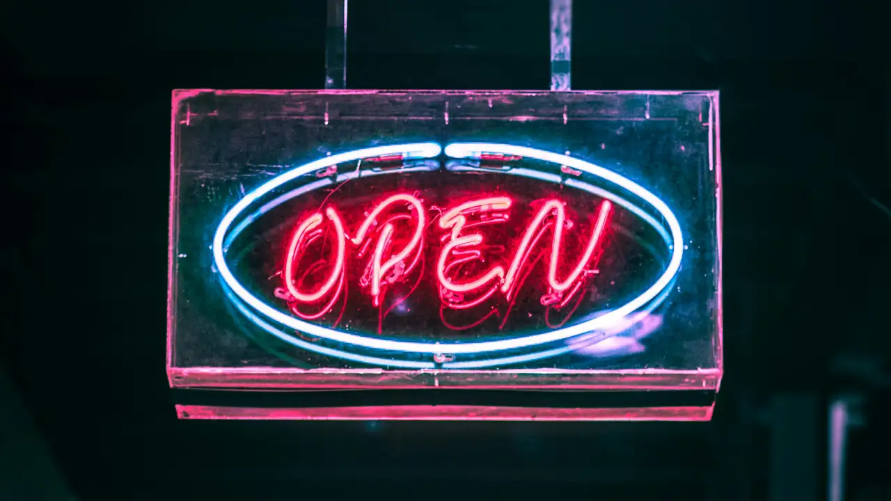 Neon sign with the word “OPEN” glowing in pink-red letters and a blue oval border on a dark background.