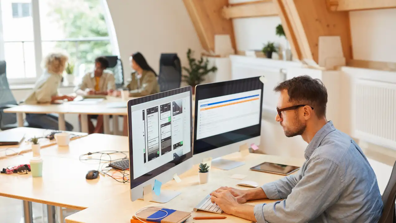 A smiling male executive in a dark suit stands at a whiteboard, pointing to a diagram and giving a presentation to a group of colleagues seated at a conference table with laptops.