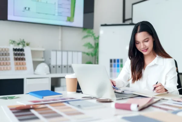 Person working at a desk with color swatches, design materials, a laptop, and office supplies in a creative studio setting.