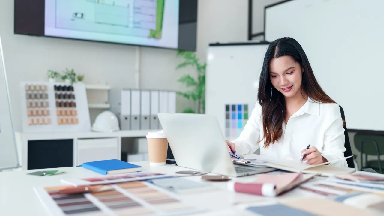 Person working at a desk with color swatches, design materials, a laptop, and office supplies in a creative studio setting.