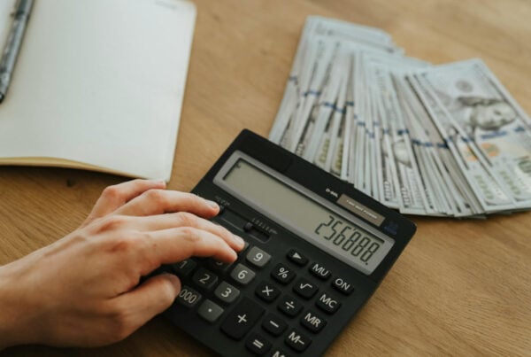 A close-up view of a hand using a calculator next to stacks of US hundred-dollar bills and an open notebook on a wooden desk.