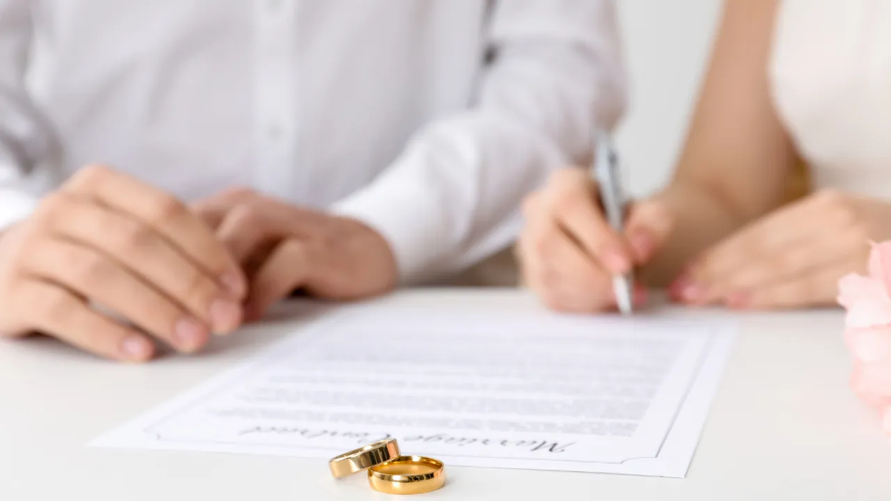 A couple sitting at a table signing a document with two gold rings placed on the paper.