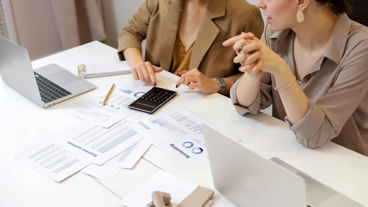 Two individuals sitting at a desk with laptops, financial documents, charts, a calculator, and office supplies during an accounting related work discussion.