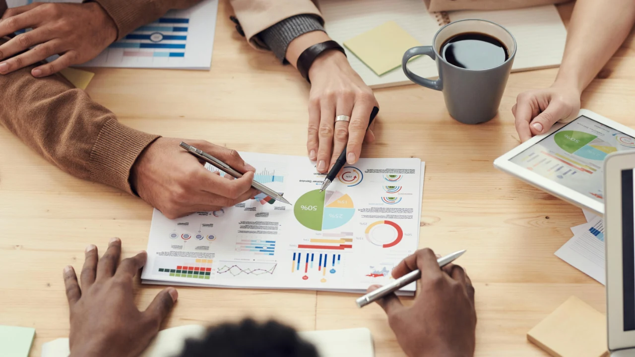 A table with charts, graphs, documents, pens, notebooks, a coffee cup, and a laptop, with several people pointing to business data on printed pages.