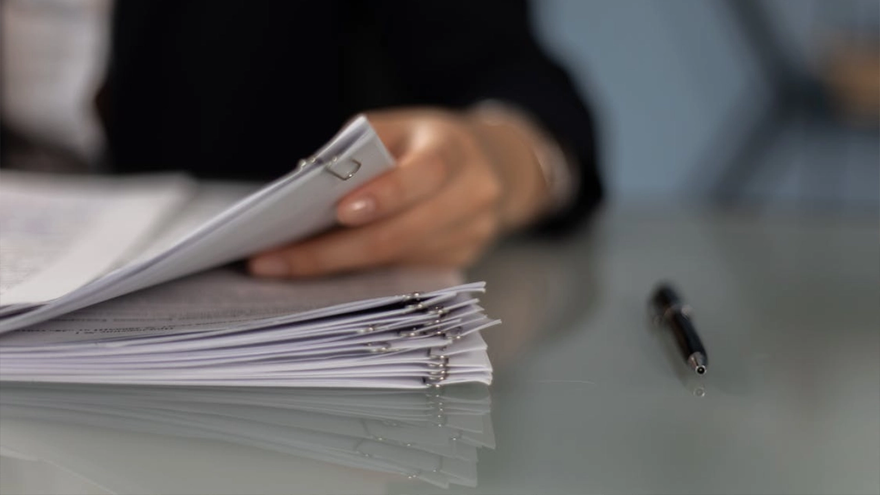 A legal professional's hand flipping through a thick stack of organized documents with binder clips on a reflective desk next to a pen.
