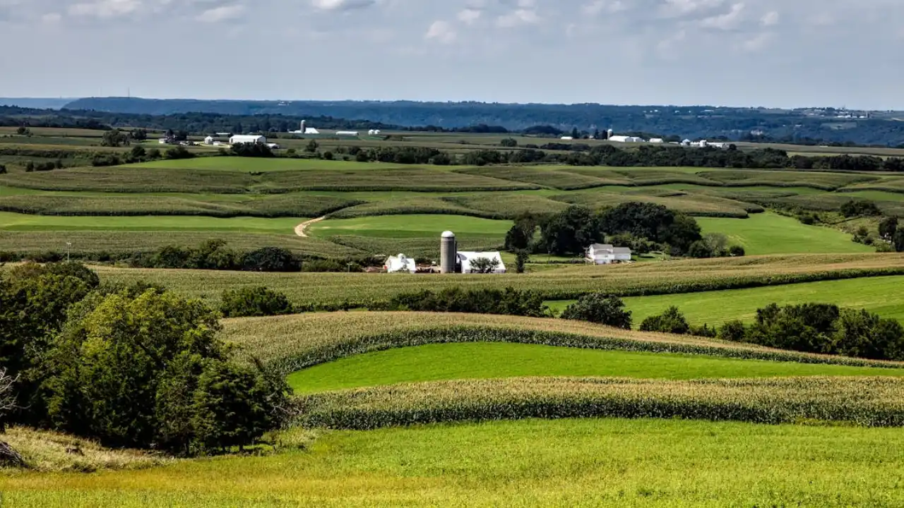 An aerial view of lush green farmland, rolling hills, and a farm with a white barn and silo under a partly cloudy sky.