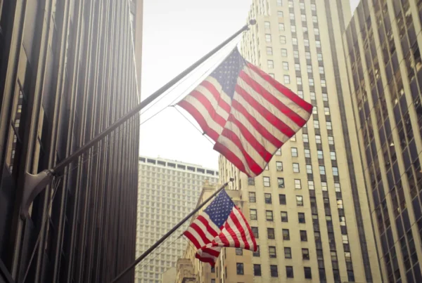 Two American flags mounted on building facades waving in front of tall, modern skyscrapers in a city business district.