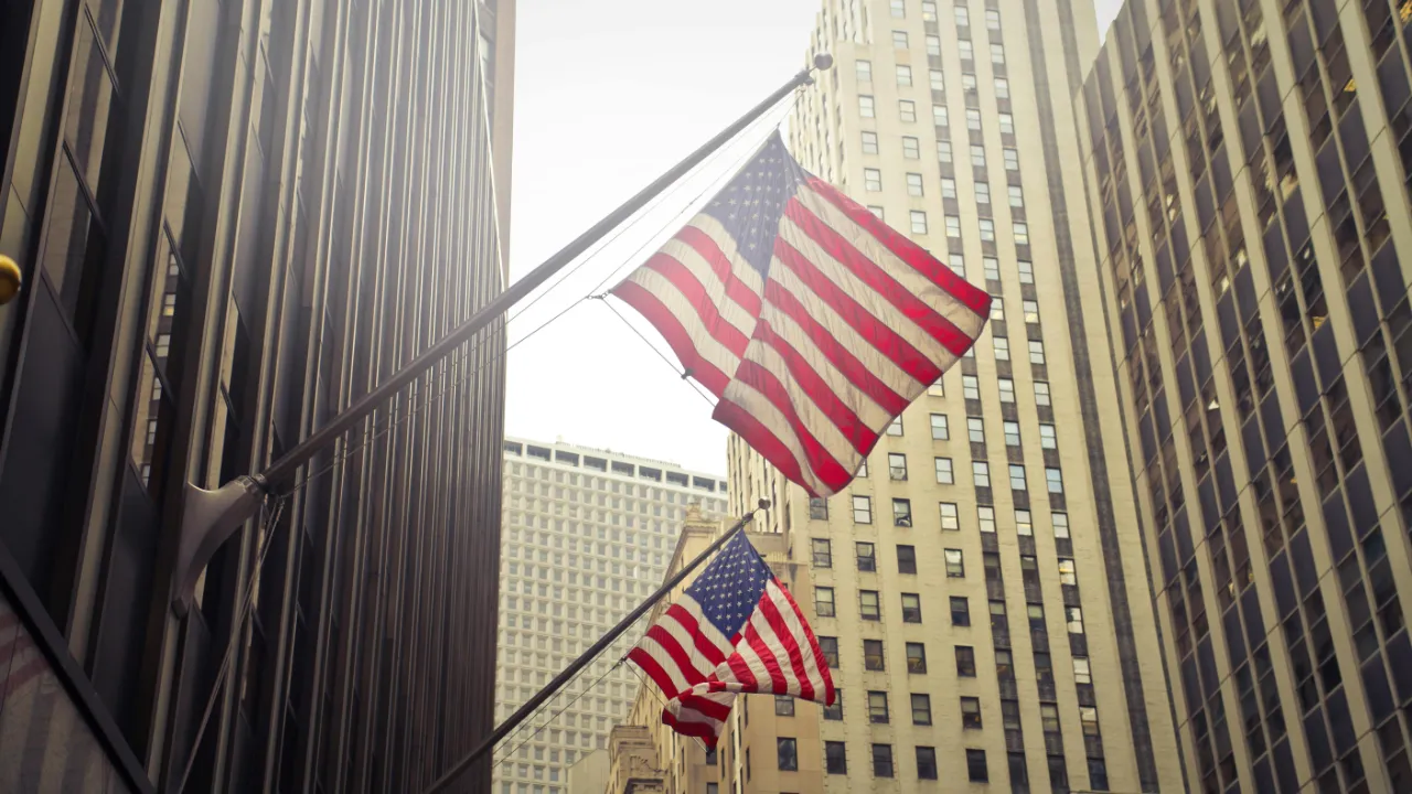 Two American flags mounted on building facades waving in front of tall, modern skyscrapers in a city business district.