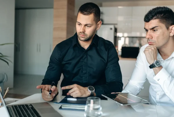 Two individuals seated at a desk reviewing a laptop, papers, and electronic devices during a business meeting.