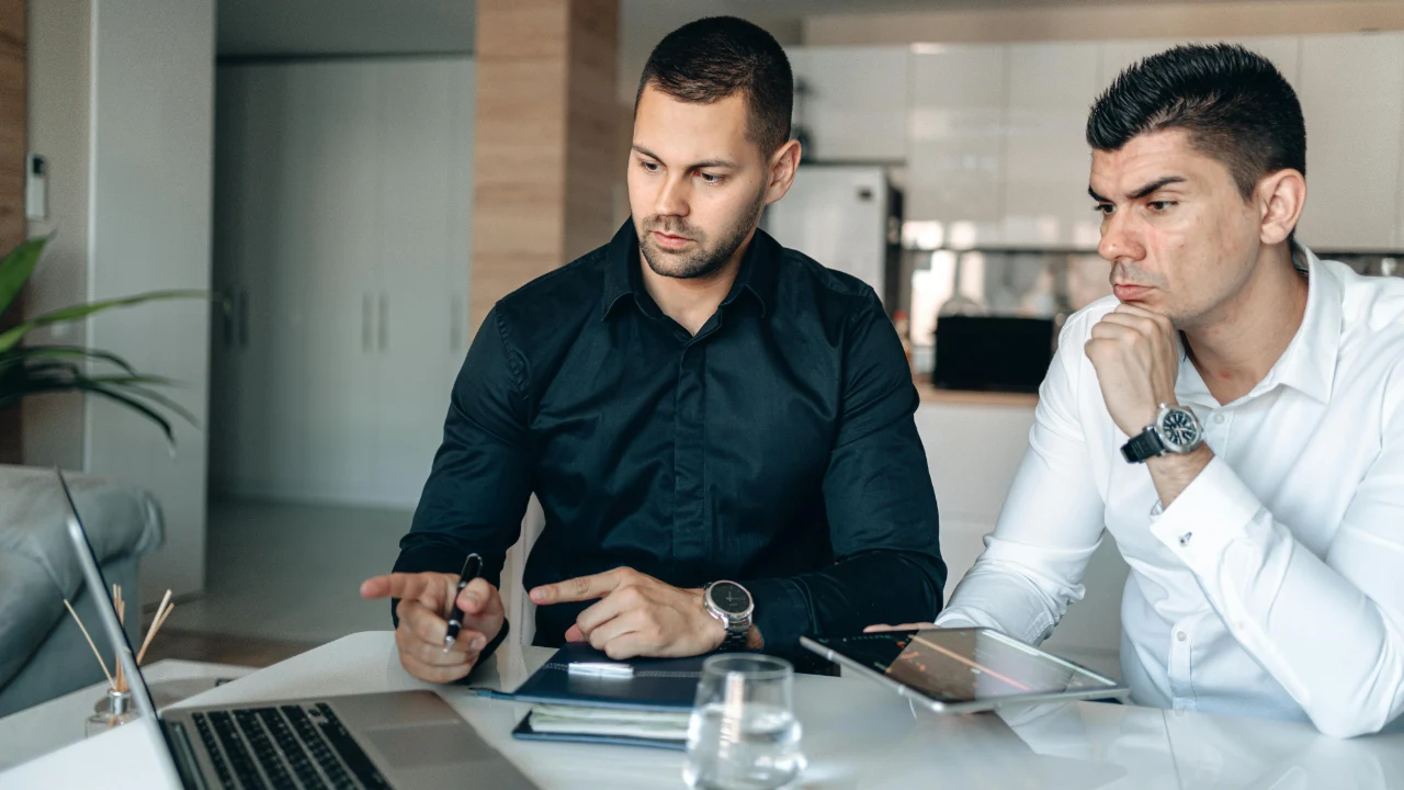 Two individuals seated at a desk reviewing a laptop, papers, and electronic devices during a business meeting.