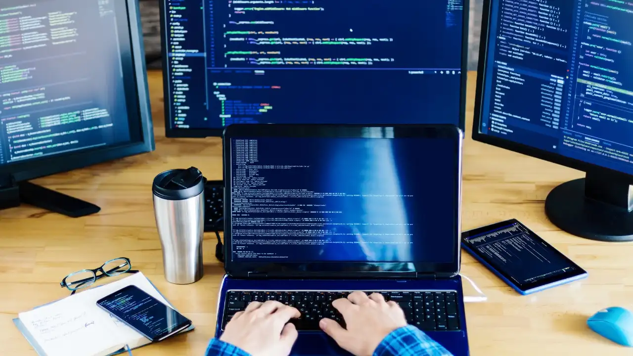 Person typing on a laptop surrounded by computer monitors displaying code.