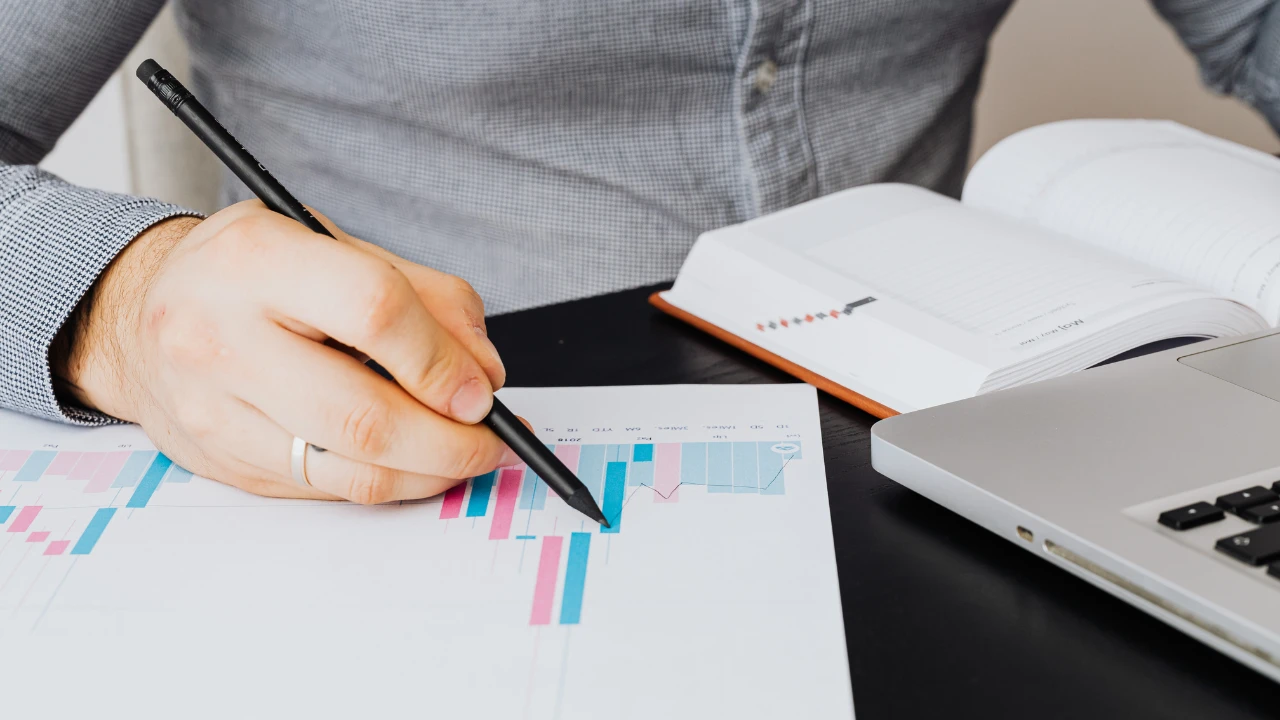 A close-up of a professional pointing a pencil at a financial bar chart on a desk with a laptop and planner.