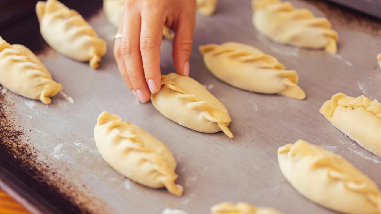 Unbaked filled pastries arranged on a metal baking tray with a hand shaping one pastry.