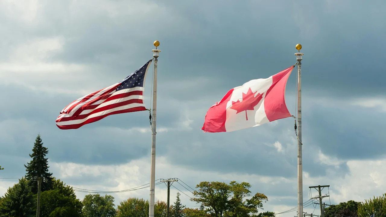 American and Canadian flags at a land border crossing under a cloudy sky.